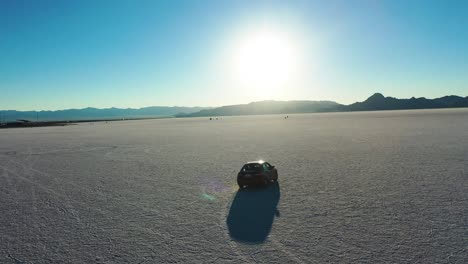 Aerial-view-of-a-red-car-driving-across-large-saltflats,-sunny-evening-in-USA