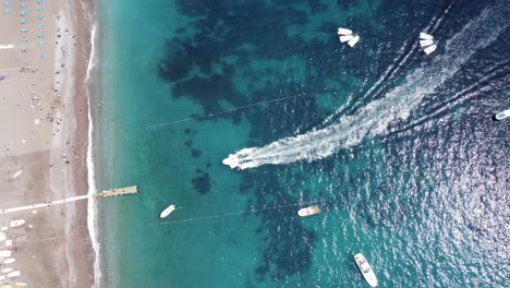 Luftaufnahme-Eines-Bootes-Von-Oben-Nach-Unten,-Das-Durch-Wunderschönes-Türkisblaues-Wasser-Fährt-Und-An-Einem-Sonnigen-Sommertag-Mit-Unberührtem-Weißem-Sandstrand-In-Positano-Ankommt