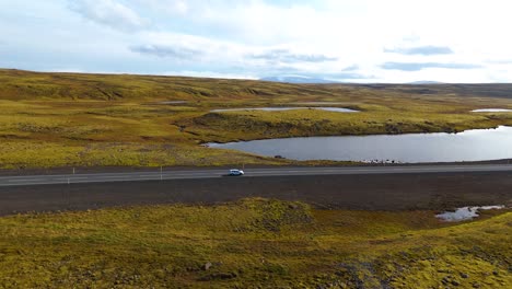 Luftaufnahme-Eines-Autos,-Das-Auf-Der-Straße-In-Einer-Malerischen-Isländischen-Landschaft-Mit-Seen-Und-Grasland-Fährt