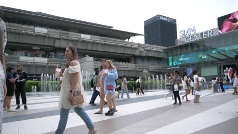 People-are-busy-walking-outside-of-Siam-Paragon-in-Bangkok-with-the-BTS-train-station-with-trains-leaving-and-stopping-for-passengers,-in-Thailand