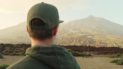 Man-wearing-green-hoodie-and-baseball-cap-looks-afar-towards-the-mountain,-handheld-closeup