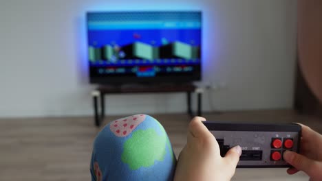 Toddler-girl-plays-with-vintage-gaming-console-during-the-evening,-moving-cinematic-shot,-TV-with-a-game-in-the-background,-static-shot