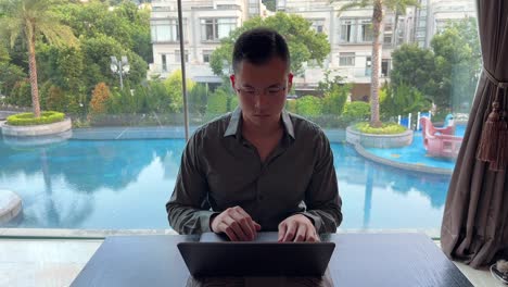 Empresario-Milenario-Asiático-Escribiendo-En-Una-Laptop-Con-Lujosa-Piscina-Y-Casas-En-El-Fondo