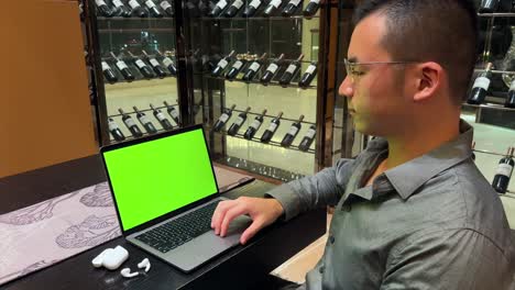 Asiatischer-Millennial-Mann-Lächelt,-Während-Er-Auf-Dem-Trackpad-Eines-Laptops-Mit-Grünem-Bildschirm-In-Einem-Luxuriösen-Zuhause-Scrollt