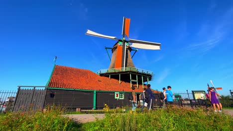 Low-Angle-Aufnahme-Einer-Windmühle,-Während-Menschen-In-Zaanse-Schans,-Niederlande,-Vorbeigehen-–-Blauer-Himmelshintergrund-Mit-Windmühle