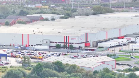 Drone-ascends-treeline-revealing-Tesco-distribution-center-with-semi-trucks-at-the-loading-dock