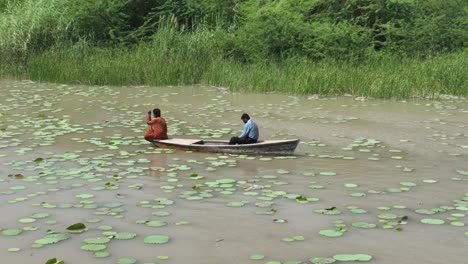 Aerial-Orbit-Motion-Around-Two-Men-On-Small-Wooden-Boat-On-Botar-Lake-With-Small-Green-Lily-Pads-Floating-On-Surface