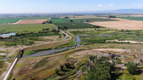 Panoramic-video-of-expansive-farmlands,-a-tranquil-river-winding-through-the-pasturelands,-and-a-drone's-journey-direct-to-the-horizon,-beneath-the-endless-blue-sky-in-Colorado
