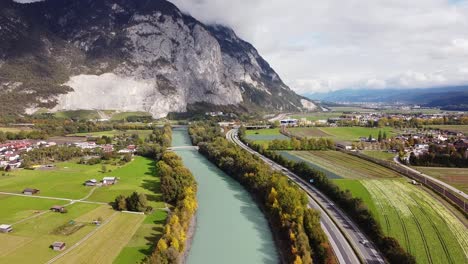 Flying-along-river-with-trees-growing-around-abd-a-street,-mountains-in-the-distance-on-a-sunny-day,-Innsbruck-Austria