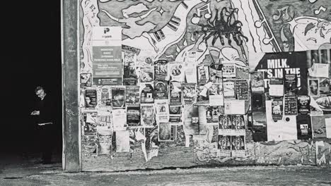 A-Old-Man-in-Black-Smoking-a-Cigarette-In-an-Ally-Checks-his-Phone-Next-to-a-Large-Downtown-Urban-Large-City-Advertising-Poster-Wall-Grimy-Hand-Painted-Street-Photography-Culture-Capture-Black-White