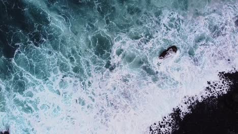 Wunderschöne-Filmische-Luftaufnahme-Von-Oben-Nach-Unten-Mit-Tiefen,-Klaren,-Blauen-Meereswellen,-Die-Zur-Schwarzen-Felsigen-Küste-Rollen