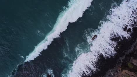 Beautiful-cinematic-top-down-aerial-of-deap-clear-blue-ocean-sea-waves-rolling-to-black-rocky-shore