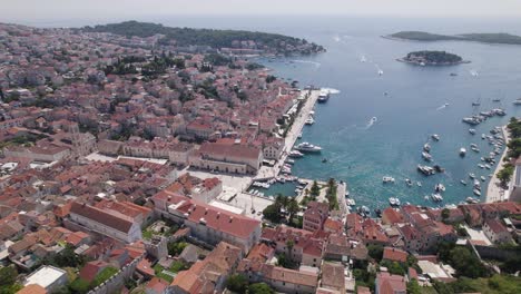 Coastal-harbor-of-Hvar-Croatia-at-midday-as-boats-enter-the-port-town,-aerial-establish