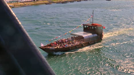 Magnífico-Barco-Con-Muchos-Turistas-Cruzando-Un-Puente-Sobre-El-Río-Duero-En-Portugal.