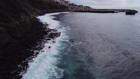 Beautiful-aerial-of-deap-clear-blue-ocean-sea-waves-rolling-to-rocky-shore,-town-on-tenerife-in-distance