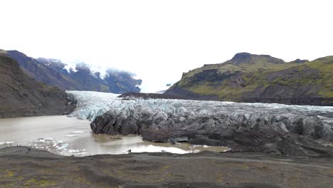 drone-shot-moving-forward-with-a-couple-of-Europeans-on-the-left-and-turning-towards-a-huge-glacier,-black-volcanic-soil-and-relief-with-little-vegetation-and-brown-water