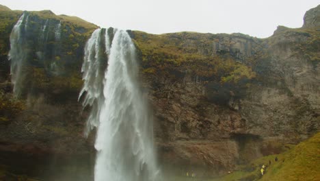 Impresionante-Inclinación-Hacia-Abajo-De-La-Cascada-De-Seljalandsfoss-Para-Turistas-Que-Caminan-Por-El-Sendero-Cerca-De-La-Montaña