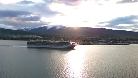 Cruise-ship-sailing-near-Icelandic-town-with-mountain-backdrop-at-sunset