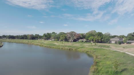 Luftdrohnenaufnahme-Einer-Radfahrerin-Mit-Einem-Kindertransporter,-Die-Auf-Dem-Grünen-Erholungspfad-Unter-Blauem-Himmel-Und-Weißen-Wolken-Im-Klaren-See,-Houston,-Texas,-Fährt