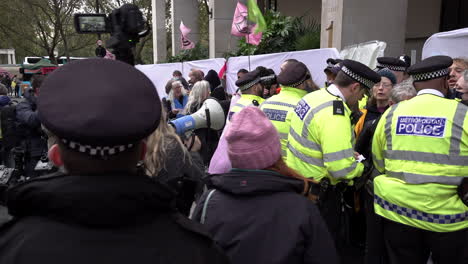 Ein-Beweissammler-Der-Stadtpolizei-Filmt-Mit-Einer-Videokamera-Auf-Einem-Einbeinstativ,-Während-Andere-Beamte-Während-Eines-Protests-Mit-Klimaaktivisten-Der-Extinction-Rebellion-Sprechen