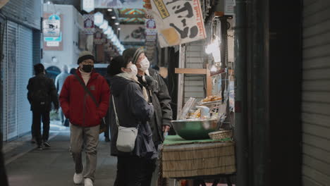 Asian-couple-in-masks-shopping-and-chatting-with-stall-vendor-in-front-of-food-stall-in-Nishiki-Market-during-the-pandemic