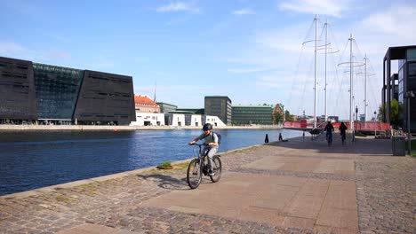 Couple-and-kid-riding-bikes-in-front-of-the-Royal-Danish-Library,-Denmark