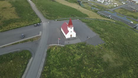 Bask-in-the-ethereal-glow-of-sunset-with-a-captivating-aerial-view-of-the-iconic-white-church-in-Vik,-Iceland,-perched-atop-a-hill-overseeing-the-town's-tranquil-panorama