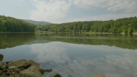 Calm-lake-with-rocky-shore-green-forest-and-sky-cloud-reflections-pan-up-shot