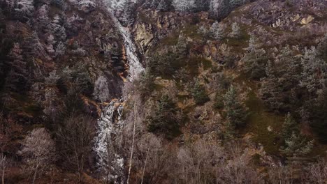 Flying-along-steep-mountain-slope-in-fall,-white-frosty-trees-with-dries-out-waterfall,-moody-drone-flying-up