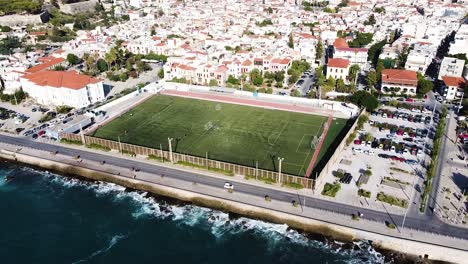 White-homes-of-Rethymno-and-green-football-field,-aerial-orbit-view