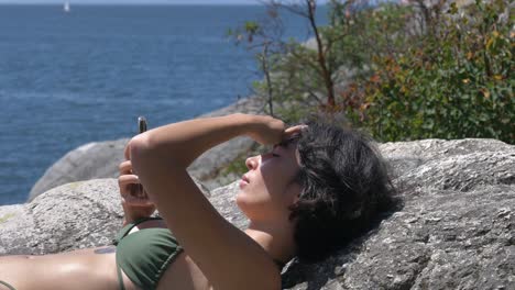 Close-up-of-beautiful-brunette-girl-in-bikini-sunbathing-on-rock-and-using-smartphone