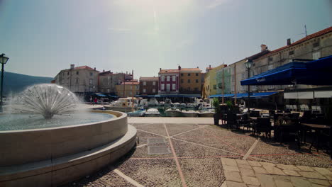 Pan-across-fountain-near-coastal-cafe-below-colorful-building-facades,-Cres-Croatia