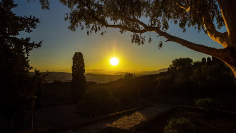 In-Malaga,-Spain,-soak-in-the-captivating-scenery-as-the-sun-rises-above-the-town's-trees,-illuminating-with-a-breathtaking-golden-hue-while-casting-an-enchanting-glow-on-the-majestic-mountain-range