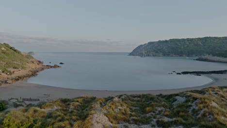 Flyover-the-sandy-shore-and-beach-of-La-Vall-towards-the-sea,-Menorca