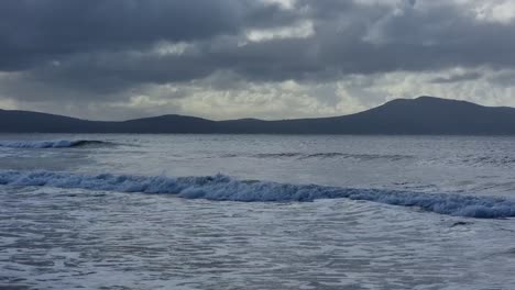 Following-rough-stormy-shimmering-waves-breaking-under-misty-Snowdonia-mountain-range-with-sunrays-shining-through-overcast-clouds