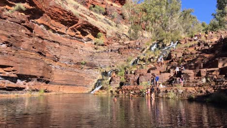 Piscina-De-Agua,-Rocas-Rojas,-Acantilados-Y-Turistas-Tomando-El-Sol-En-Las-Cataratas-Fortescue-En-Karijini,-Australia-Occidental