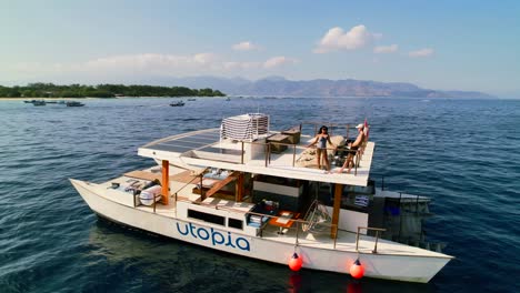 Junge-Paarreisende-Genießen-Den-Blick-Von-Der-Dachterrasse-Auf-Die-Küste-Von-Gili-Von-Einer-Luxuriösen-Katamaran-Yacht-Aus,-Die-Im-Blauen-Meer-Kreuzt-–-Luftparallaxenansicht