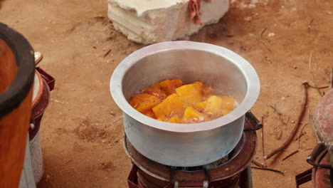 Black-hand-is-stirring-large-chunks-of-yellow-orange-pumpkin-in-a-metal-pot,-which-is-placed-on-a-small,-cylindrical-metal-stove