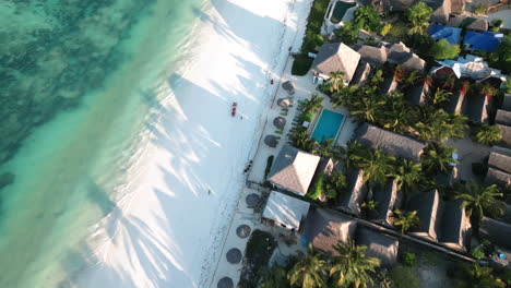 Aerial-shot-of-a-white-sandy-beach-in-Zanzibar-with-thatched-roof-structures-and-palm-trees,-indicative-of-a-resort-area-beside-the-clear-blue-ocean