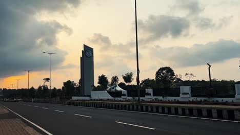 Monument-with-clock-on-Soraba-Avenue-during-sunset,-biker-passing-by,-India