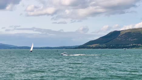 Weißes-Schnellboot-Und-Segelboot-Fahren-Inmitten-Der-Atemberaubenden-Schönheit-Eines-Sonnigen-Tages-Mit-Wolken,-Eingerahmt-Von-Den-Majestätischen-Mourne-Mountains-In-Nordirland