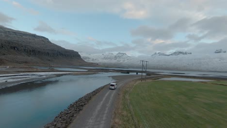 Drone-shot-of-a-white-van-driving-on-a-long-road-surrounded-with-lawns,-water-and-the-mountains,-in-Iceland