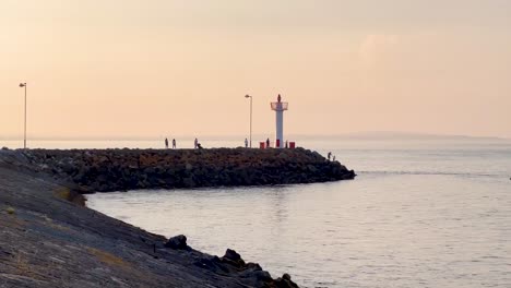 Golden-Hour-Serenity:-People-Walking-to-Howth-Lighthouse-with-Calm-Waves,-Ireland