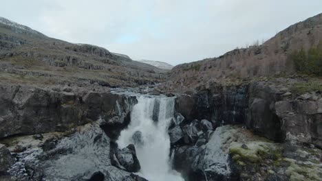 Drone-Volando-Sobre-Una-Hermosa-Y-Amplia-Cascada,-Hacia-Un-Río-Lleno-De-Rocas-En-Islandia