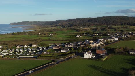 Aerial-view-flying-above-Anglesey-small-farming-village-vibrant-green-agricultural-farmland-and-glowing-sunset-skyline