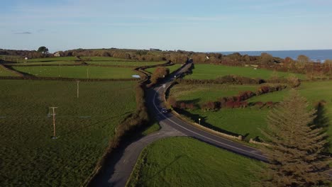 Aerial-view-across-quiet-curving-Anglesey-coastal-road-surrounded-by-lush-vibrant-agricultural-farmland-at-sunset
