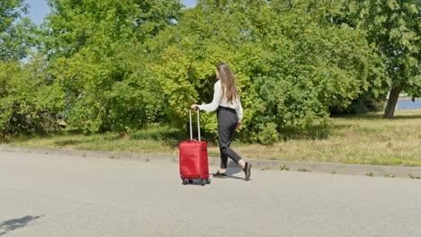 Girl-walking-through-green-park-in-slow-motion-with-red-luggage,-Riga