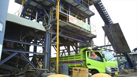 Transport-truck-driver-inspects-hotmix-asphalt-falling-from-the-mixer-into-the-truck-bed-at-the-asphalt-mixing-plant-to-be-transported-or-supplied-to-the-project-site-in-road-construction