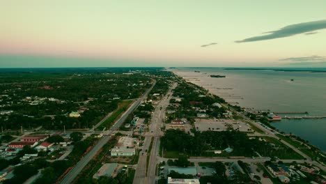 Aerial-shot-of-Sebastian,-Florida-showcasing-a-busy-road,-adjacent-shoreline,-and-expansive-greenery