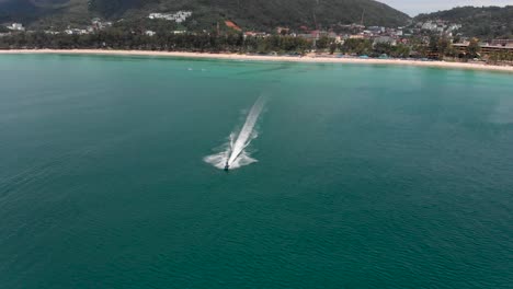 Hovering-in-position-and-slowly-tilting-the-drone-from-a-panoramic-view-of-Kata-beach-to-focus-on-a-speeding-jetski-in-Kata-Beach